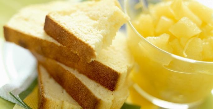 Fotografia em tons de amarelo, em uma bancada de madeira de cor branca. Ao centro, um prato verde contendo as fatias de pão com um recipiente ao lado contendo alguns pedaços de frutas.