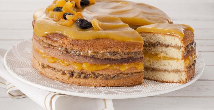 Fotografia em tons de amarelo em uma bancada de madeira branca com um pano branco com listras douradas e ao centro um prato branco com o bolo de coco com recheio de doce de leite com ameixa e geleia de damasco em cima.