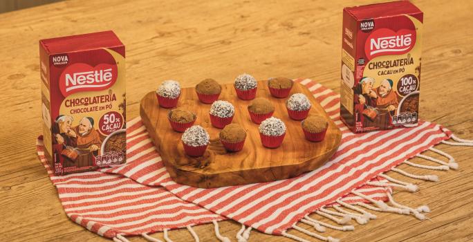 Fotografia em tons de vermelho em uma bancada de madeira de cor marrom. Ao centro, um pano branco com listras vermelhas com uma tábua de madeira em cima contendo os brigadeiros. Ao lado, há duas caixinhas com cacau e chocolate em pó dois frades.