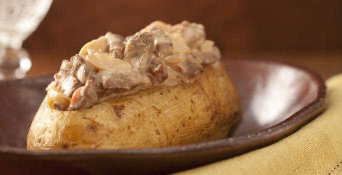 Fotografia em tons de marrom e pardo, em uma bancada contém um pano cor pardo com um garfo por cima, um recipiente marrom com uma batata com pedaços de carne com molho por cima e ao lado um copo transparente com agua