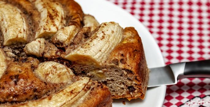 Fotografia em tons de amarelo, em uma bancada de madeira com uma toalha quadriculada vermelha. Ao centro, um prato redondo branco contendo o bolo de banana.