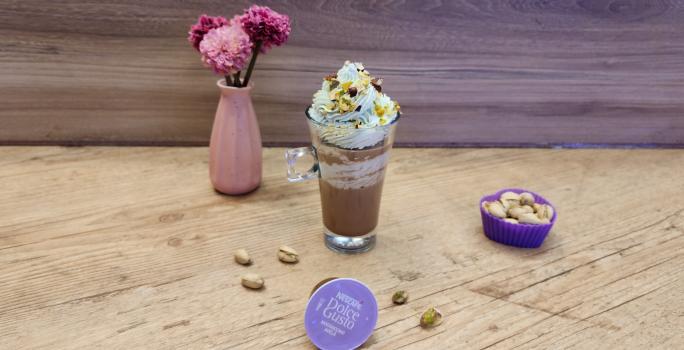 Fotografia em tons de lilás em uma bancada de madeira, com uma xícara de vidro e o Mochaccino Avelã com chantilly de pistache em cima para decorar. Em destaque, a cápsula de Dolce Gusto e ao fundo um vaso rosa com flores rosas.