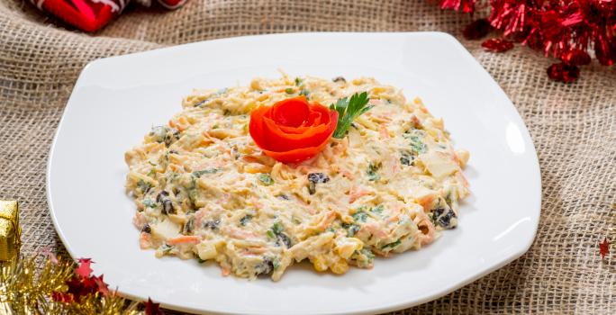 Fotografia de um salpicão tradicional cremoso com frango, cenoura, salsinha e uva passa em um prato branco quadrado. Por cima da salada, um tomate em formato de rosa e ao lado, uma folha de salsinha. Ao redor, decorações de Natal.