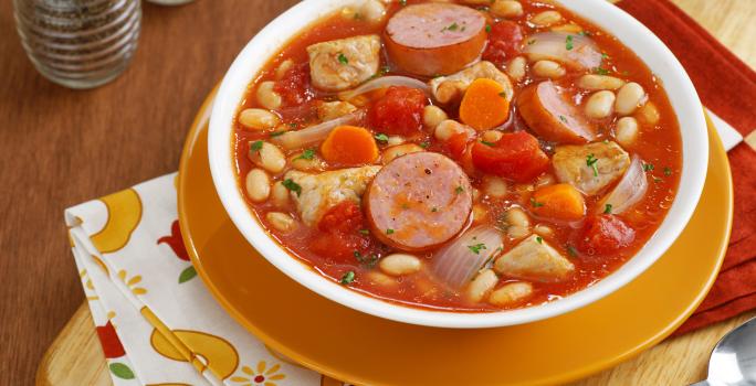 fotografia em tons de laranja e vermelho de uma bancada marrom vista de cima, contém um prato redondo laranja com um recipiente redondo branco que contém Cassoulet com pedaços de linguiça, feijão branco e abóbora.