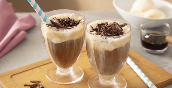 Foto em tons de marrom da receita de affogato de nescafé dolce gusto servida em duas xícaras de vidro dispostas sobre uma tábua de madeira com raspas de chocolate ao canto, além de canudos listrados em azul. Ao fundo uma xícara de café e sorvete.