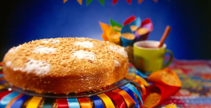 Fotografia em tons coloridos em uma bancada com uma toalha junina, um bolo redondo em cima de um prato com fitas de cetim decorando ele e bandeirolas de festa junina ao fundo.