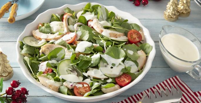 Foto em tons claros da receita de salada de chester com molho de iogurte servida em um bowl grande de porcelana branca com uma xícara de iogurte ao lado sobre uma mesa de madeira azul