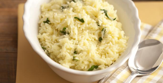 Fotografia em tons de amarelo em uma bancada de madeira com um pano listrado em em bege e amarelo e um prato fundo branco com o arroz com rúcula.