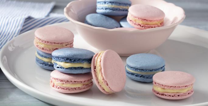 Fotografia em tons de azul e rosa em uma bancada de madeira branca clara com vários macarons coloridos em um prato branco e um paninho azul listrado.