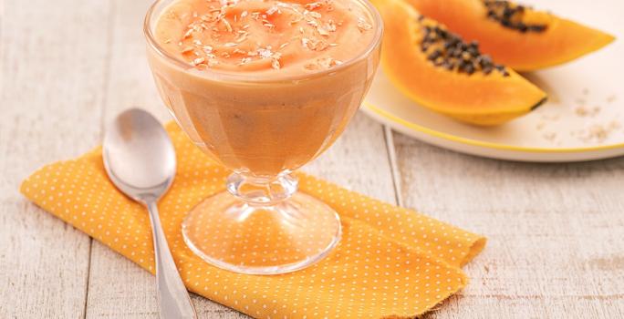 Foto em tons de laranja da receita de creme de papaia servida em uma taça de vidro sobre um pano laranja em uma mesa de madeira. Ao lado há um colher prateada e ao fundo há um prato com duas fatias médias de mamão