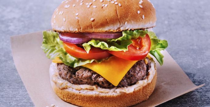 Fotografia com um fundo cinza, um pedaço de papel manteiga cortado em quadrado. Sobre ele, um hambúrguer com carne bovina, uma fatia de queijo, alface, duas rodelas de tomate, cebola roxa e um molho na base do lanche.