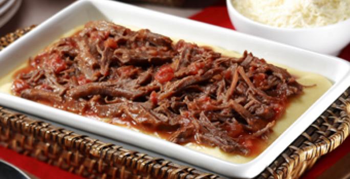 Fotografia em tons de vermelho, branco e marrom, com refratário sobre bandeja de vime com carne desfiada e molho de tomate, sobre guardanapo vermelho, ladeada por pote branco com arroz e uma colher, colher de servir, pratos, tudo sobre bancada de madeira.