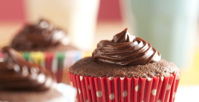 Fotografia em tons de branco e vermelho de uma bancada branca com bolinhas coloridas, sobre ela três cupcakes de Nescau. Ao fundo dois copos com canudos coloridos.