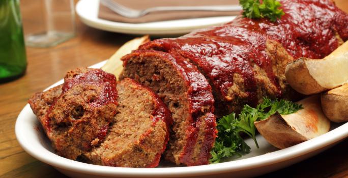 Fotografia de carne com aveia ao forno, metade cortada em fatias e dourada por cima dentro de um refratário branco oval. Ao lado, algumas batatas assadas, e ao fundo, um prato branco com um garfo encima, sobre uma mesa de madeira.