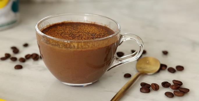 Foto aproximada de uma receita de café cremoso servido em uma xícara de vidro, com uma colher dourada ao lado e alguns grãos de café espalhados na mesa