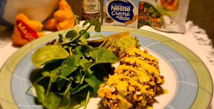 Foto aproximada de um prato redondo com bordas verde e azul com a receita de frango empanado servido com agrião. Ao fundo há produtos Maggi e Nestlé Creme de Leite e uma parte de uma pelúcia da Galinha Azul