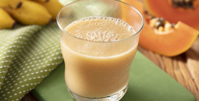 Fotografia em tons de laranja e verde de uma bancada de madeira com um suporte verde e um paninho verde com bolinhas brancas, sobre ele um copo de vidro com a vitamina de mamão e banana. Ao fundo uma fatia de mamão e um cacho de banana.