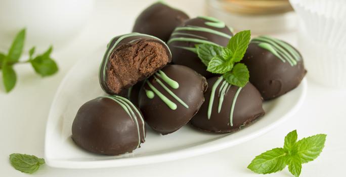 Fotografia em tons de branco, marrom e verde de uma bancada branca vista de frente, contém um prato branco com bombons de chocolate e folhas de hortelã em volta para decorar.