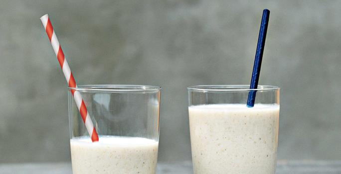 Fotografia em tons de cinza e branco de uma bancada cinza sobre ela dois copos de vidro com vitamina de banana e dois canudos, um azul e um listrado vermelho e branco.