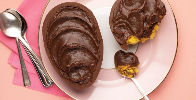 Fotografia em tons de rosa em uma bancada de madeira rosa, um pano rosa, um prato rosa redondo com o ovo de Páscoa de colher de bolo de cenoura recheado com brigadeiro.