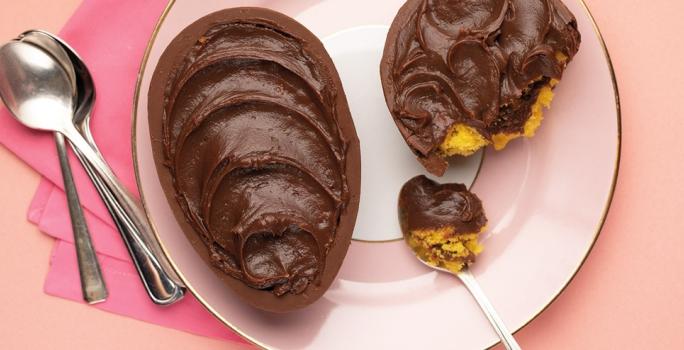 Fotografia em tons de rosa em uma bancada de madeira rosa, um pano rosa, um prato rosa redondo com o ovo de Páscoa de colher de bolo de cenoura recheado com brigadeiro.