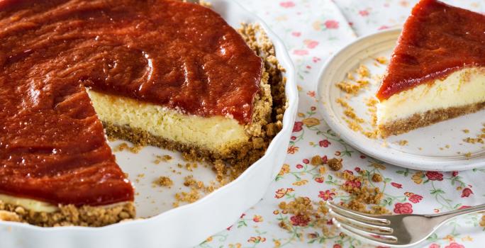 Fotografia em tons de vermelho em uma mesa com uma toalha branca com flores coloridas, um recipiente redondo raso com a cheesecake dentro dele. Ao lado, um prato raso branco com uma fatia da cheesecake.