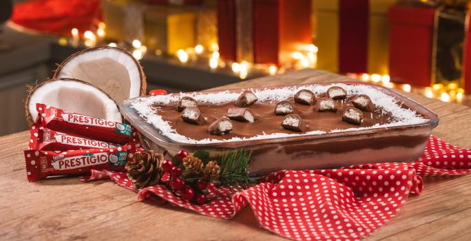 Foto da receita de Pavê de Prestígio. Observa-se o pavê em um recipiente refratário decorado com Prestígios cortados em cima. Atrás, presentes e luzes de Natal decoram a foto.