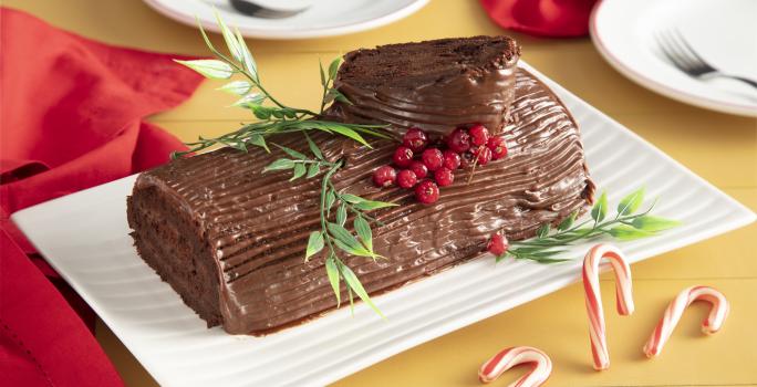 Fotografia em tons de vermelho em uma bancada de madeira amarela, um pano vermelho ao lado e ao centro, um recipiente retangular branco com o rocambole de chocolate em cima com decoração natalina.