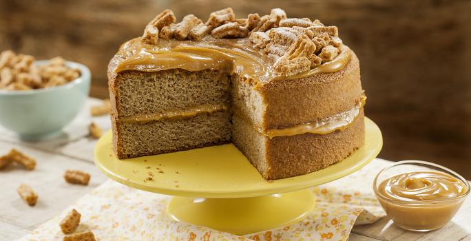 Fotografia em tons de amarelo em uma mesa de madeira com um pano bege com flores laranja e amarela, um suporte amarelo com o bolo de Moça Churros. Ao lado, um potinho com doce de leite. Ao fundo, um potinho azul com churros pequenos.