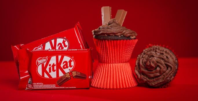 Fotografia em tons de vermelho em uma bancada de madeira de cor vermelha. Ao centro, um cupcake em pé em cima de uma forminha. AO lado direito, um cupcake deitado e ao lado esquerdo, 2 embalagens de kitkat ao laite.
