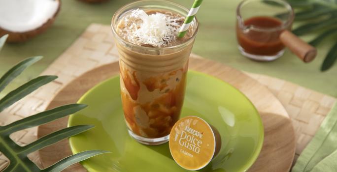 Foto da receita de iced coffee coco com caramelo servida em um copo de vidro alto com um canudo listrado verde em cima de um prato verde sobre uma tábua de madeira