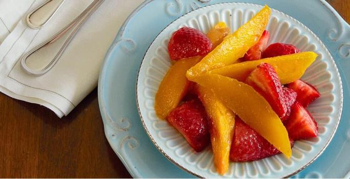 fotografia em tons de marrom, azul, amarelo e vermelho de uma bancada marrom vista de cima, contém um prato azul redondo com um pratinho redondo branco e dentro pedaços de frutas.