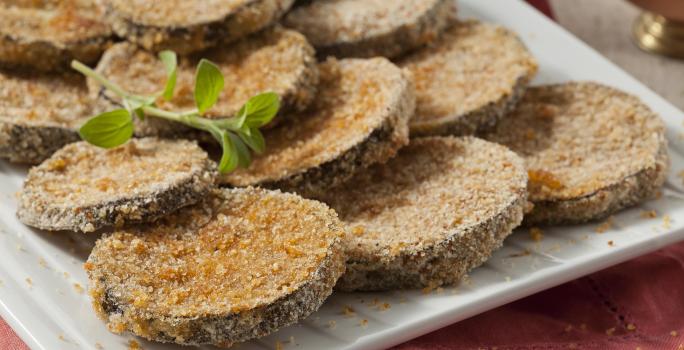 Fotografia em tons de vermelho em uma bancada de madeira com um pano vermelho, um recipiente branco retangular com as berinjelas à milanesa prontas.