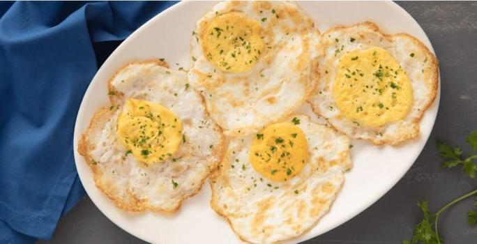 Fotografia em tons de amarelo em uma bancada de madeira escura, com um pano azul ao lado, um prato oval branco com quatro ovos fritos bem cozidos.