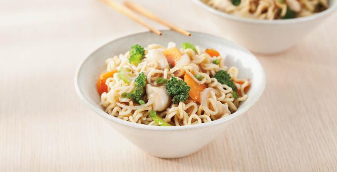 Foto de uma tigela branca com yakissoba de frango dentro e encostada nela há dois hashis. No fundo há outra tigela também com Yakissoba