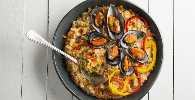 Fotografia em tons de branco e preto de uma bancada branca, sobre ele um prato preto com arroz com frutos do mar e uma colher.