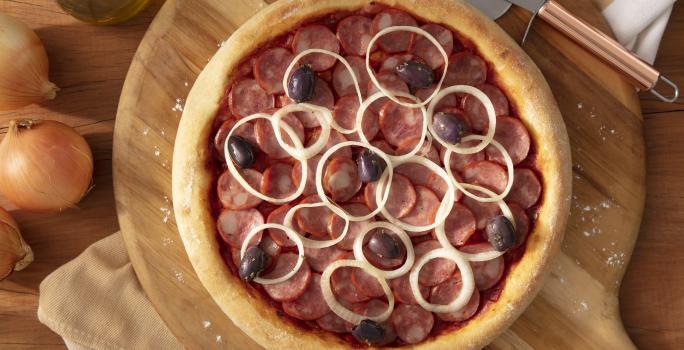 Fotografia em tons de marrom em uma bancada de madeira, uma tábua de madeira redonda com a pizza de calabresa em cima dela. Ao lado, cebola, potinho de azeite e cortador de pizza.