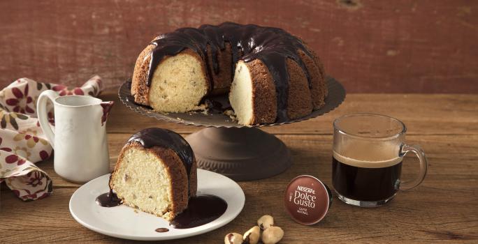 Fotografia em tons de marrom de uma bancada vista de frente, contém um pratinho redondo branco com uma fatia de bolo, um suporte para servir bolo com um bolo com cobertura e ao lado uma xicara transparente com café