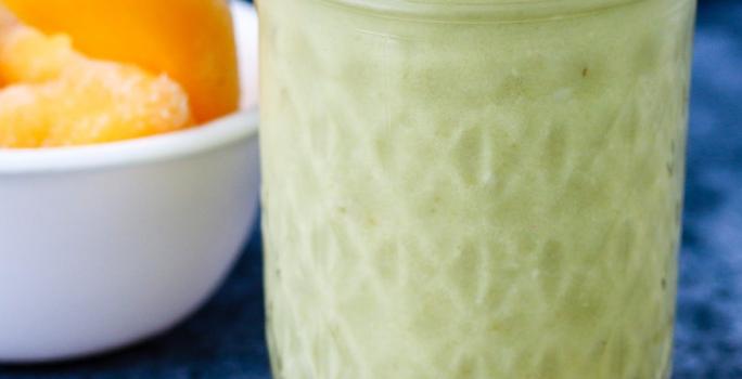 Fotografia em tons de verde em uma mesa de madeira com uma toalha azul de veludo. Ao centro, um copo grande de vidro com o smoothie de matchá e pêssego. Ao lado, uma colher dourada e um potinho com pedaços de pêssego.