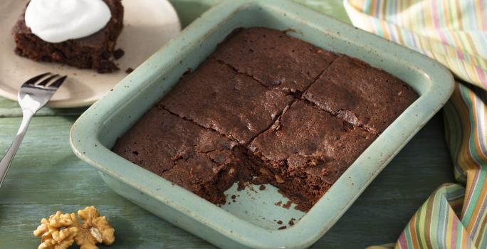 Fotografia em tons de verde em uma bancada de madeira verde, um recipiente verde quadrado com o brownie de chocolate com nozes e um pano colorido ao lado. Ao fundo, um prato redondo bege com uma fatia do brownie.