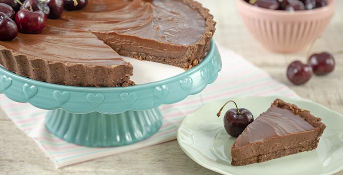 Fotografia em tons de marrom, branco, azul e rosa de uma bancada cinza vista de frente. Um recipiente azul contém uma torta de chocolate com varias cerejas e com uma fatia retirada, ao lado um prato branco com a fatia de torta e uma cereja do lado.