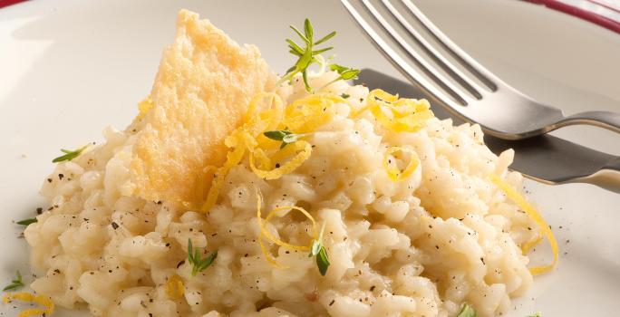 Fotografia de um prato branco com uma listra vermelha na borda, onde está servido o risoto decorando com raspas de limão siciliano e uma crosta crocante de queijo parmesão. Ao fundo uma taça de vidro transparente e um guardanapo de tecido branco.