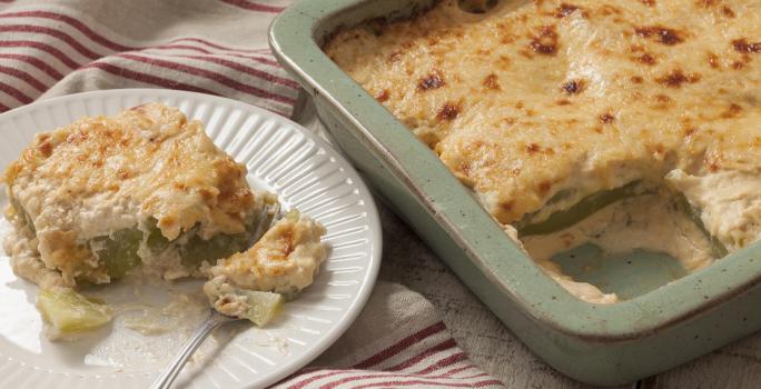 Fotografia em tons de branco e dourado, forma com Chuchu Gratinado sobre bancada de madeira rústica e prato branco com um garfo e uma porção do Chuchu Gratinado sobre guardanapo branco com listras de cor vinho.