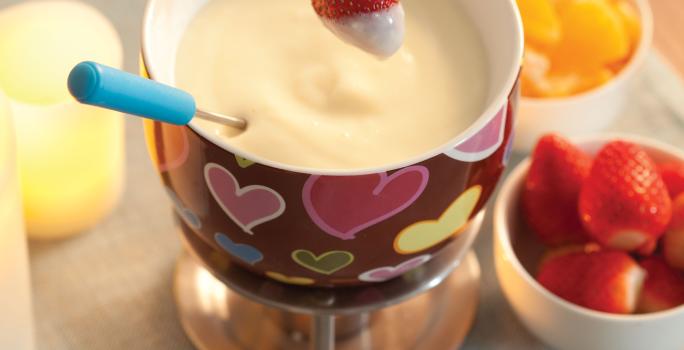 Fotografia em tons de cinza, vermelho e amarelo, com aparelho para fondue com fondue de chocolate branco com morango suspenso, no lado direito potinho com morango e outro com frutas cortadas, decoração amarela do lado esquerdo, tudo sobre toalha cinza.