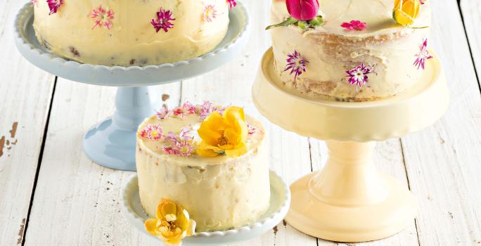 Fotografia em tons de branco, azul e pardo de uma bancada de madeira cinza vista de cima. 3 pratos para bolo em diferentes tamanhos com bolos encima e cada bolo é decorado com flores rosas e amarelas.