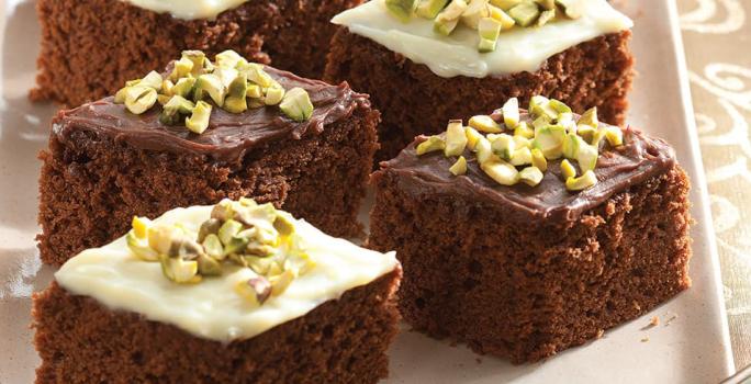 Fotografia em tons de marrom em uma mesa de madeira com uma toalha de renda, um recipiente branco retangular com os bolos de chocolate com cobertura e pistache.