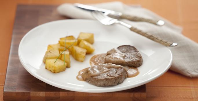 Fotografia em tons de branco e marrom de uma mesa de madeira, vista de lado, ao centro um prato redondo branco, com o filé regado com molho e batatas assadas para acompanhar. Ao lado talheres e um guardanapo de linho em tons beges.