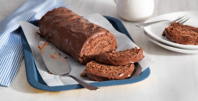 Fotografia em tons de azul, branco e marrom, ao centro da foto o rocambole todo de chocolate com 2 fatias na bandeja azul, ao lado está um prato branco com uma fatia do rocambole e do outro um guardanapo listrado de azul e branco.