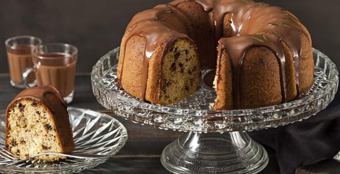 Fotografia em tons de marrom e preto de uma bancada preta vista de frente. Contém um prato redondo transparente com uma fatia de bolo com calda de chocolate por cima, um recipiente transparente para servir o bolo com o bolo sem uma fatia e calda por cima.