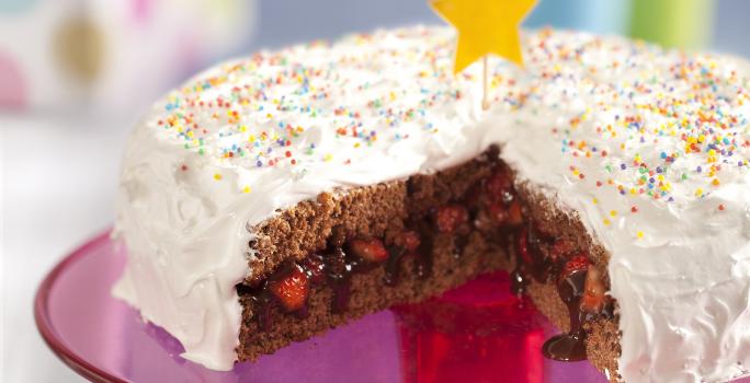 Fotografia em tons de rosa em uma bancada cinza com um suporte para bolo central na cor rosa com o bolo de aniversário confeitado em cima com cobertura branca e confeitos coloridos, cortado ao meio e recheado com chocolate e morango.